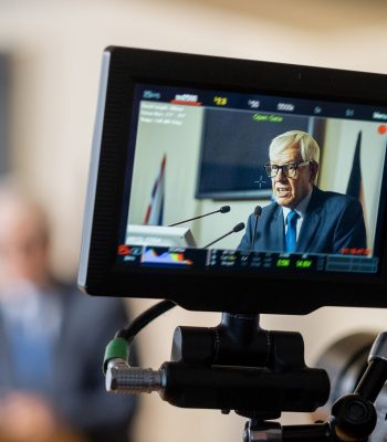 Display of camera with aged male delegate making report at political conference or some other event
