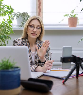 Mature confident business woman working remotely at home, using laptop smartphone for video communication and stream recording. Female consultant, psychologist, teacher, blogger, lawyer working online